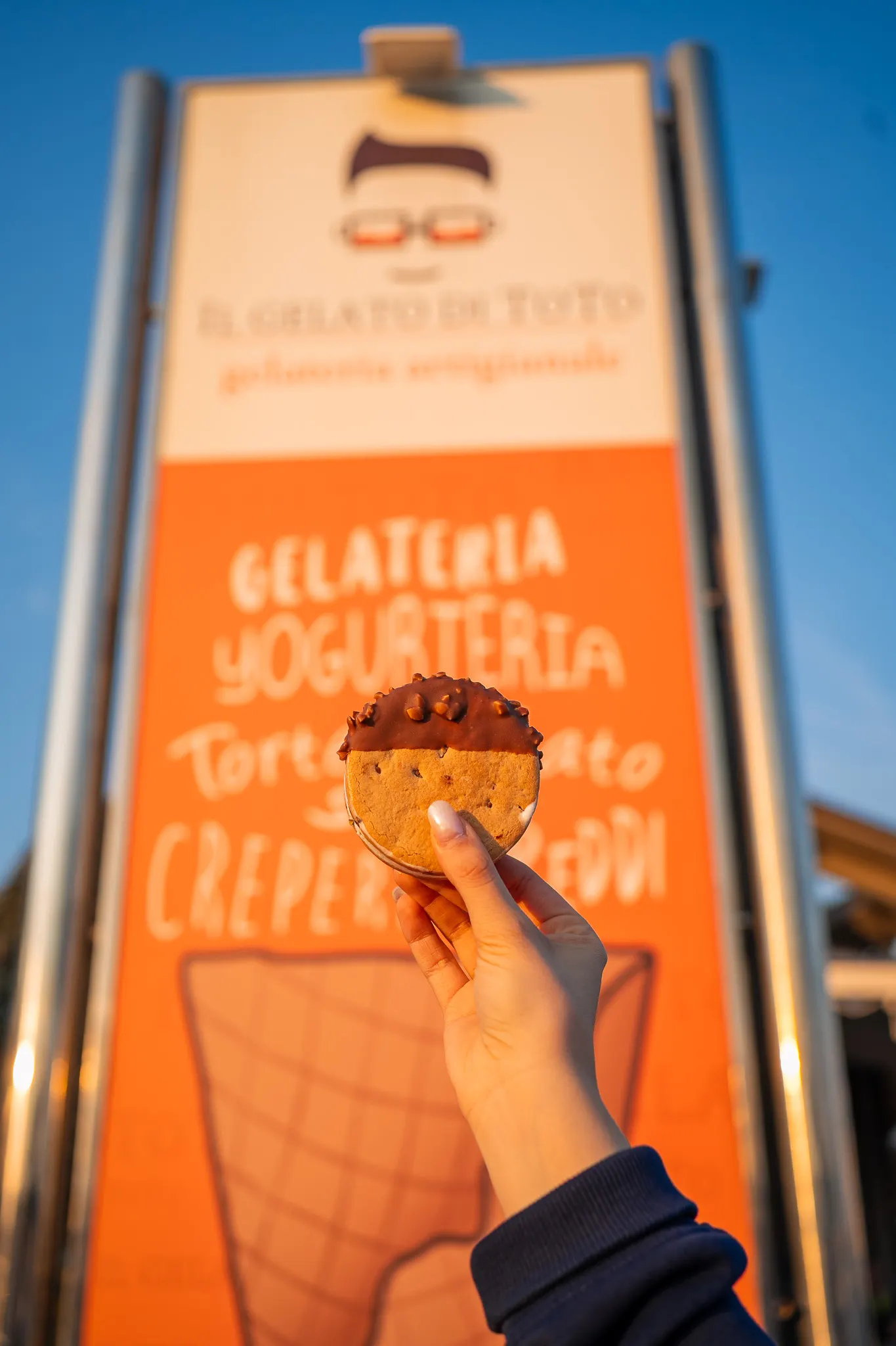 Una mano solleva il biscotto gelato al cioccolato davanti al cavalletto della gelateria