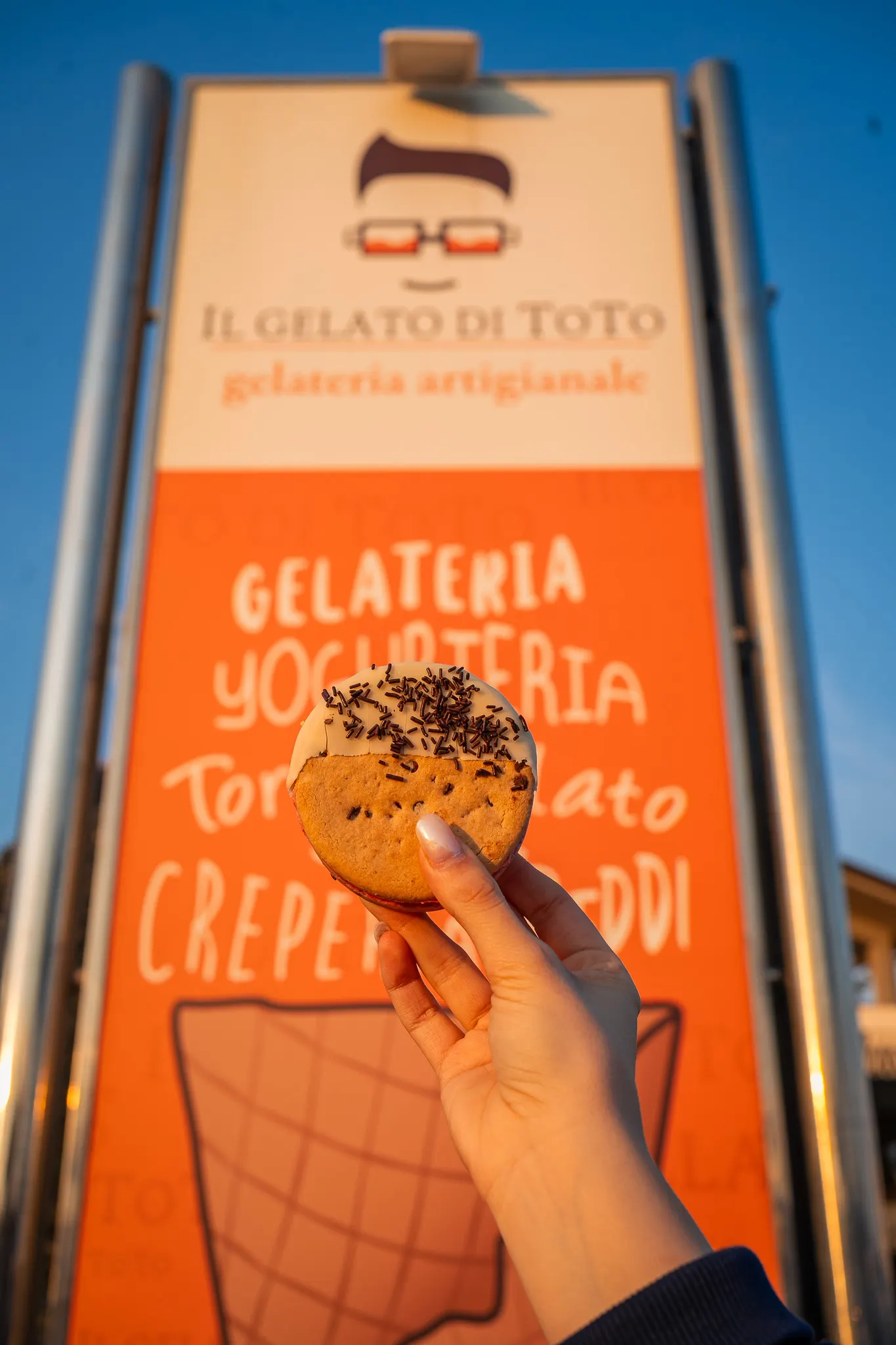 Un biscotto gelato al cioccolato davanti all'insegna arancione della gelateria, al tramonto